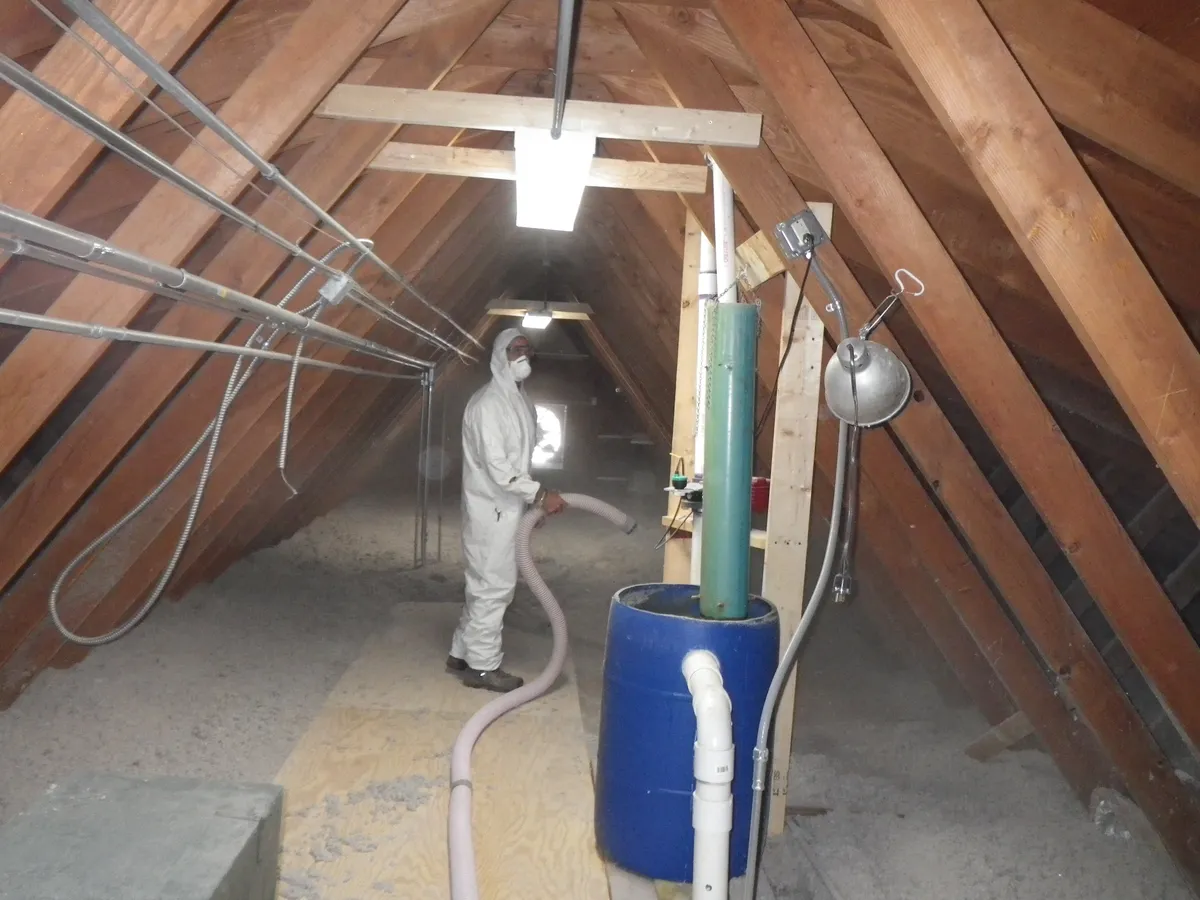 Worker in a white protective suit and mask blowing loose cellulose insulation through a hose across an attic floor under wooden rafters