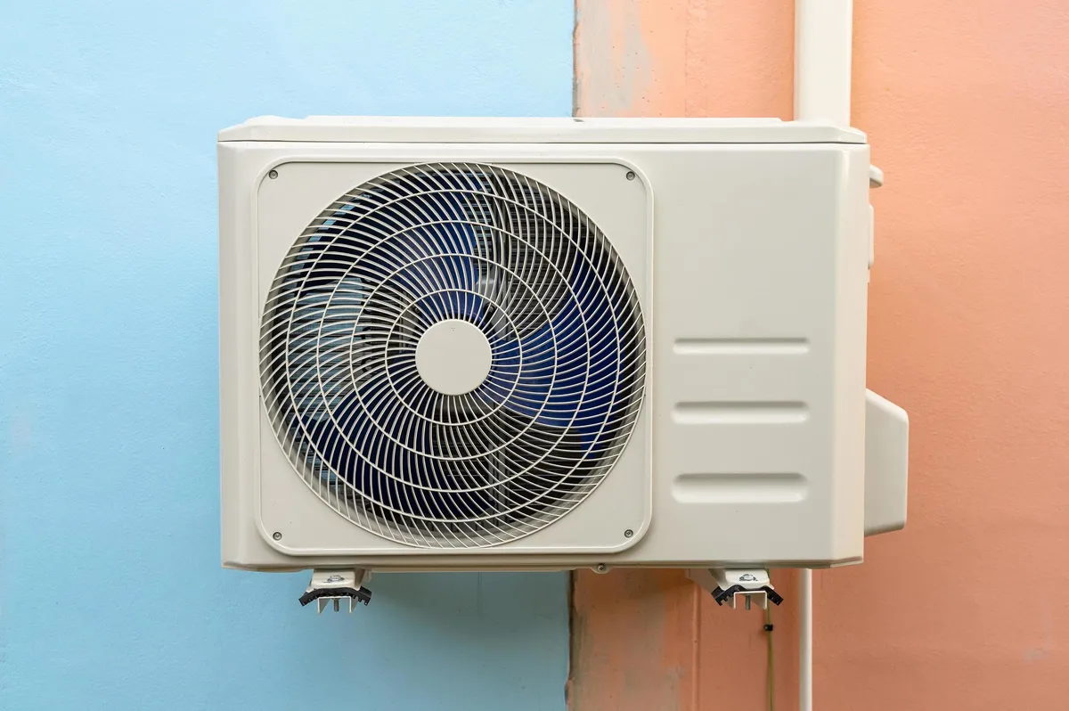 White outdoor unit of a ductless system mounted on a blue and peach wall, with a large round fan grille in the center