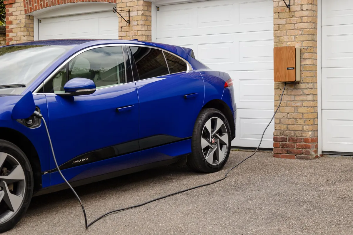 Blue electric car plugged into a wall-mounted home charger beside a brick garage, with the charging cable on the driveway