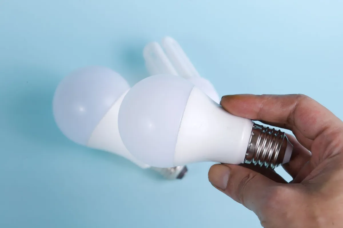 Hand holding an LED light bulb in front of other light bulbs on a pale blue background
