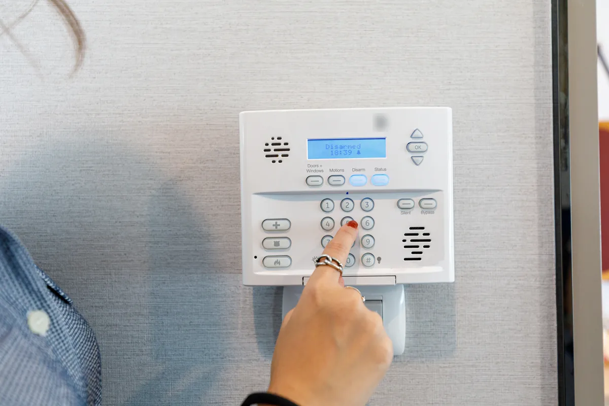 A hand pressing a number on a wall-mounted home alarm keypad whose display reads Disarmed 18:39