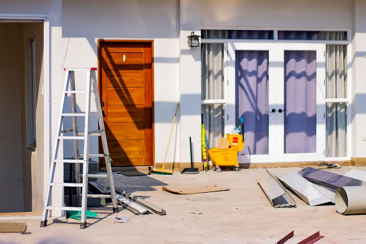 Aluminum ladder, mop bucket, broom and sheets of metal flashing on the ground outside the entrance of a white house under repair