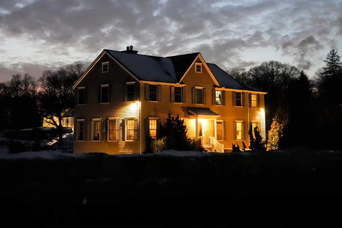Two-story house with warmly lit windows at dusk, snow on the ground and a clouded winter sky