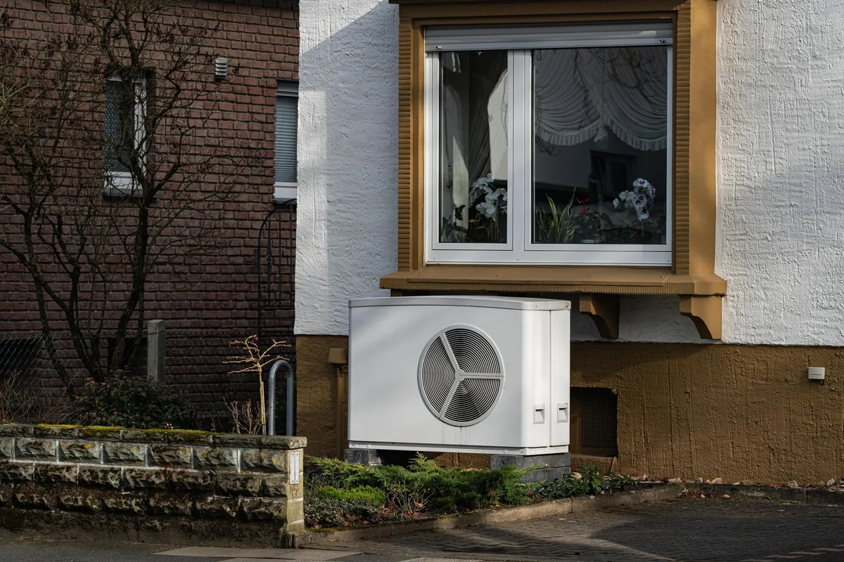 White outdoor heat pump unit installed in front of a house wall, under a window with orchids, next to a brick wall