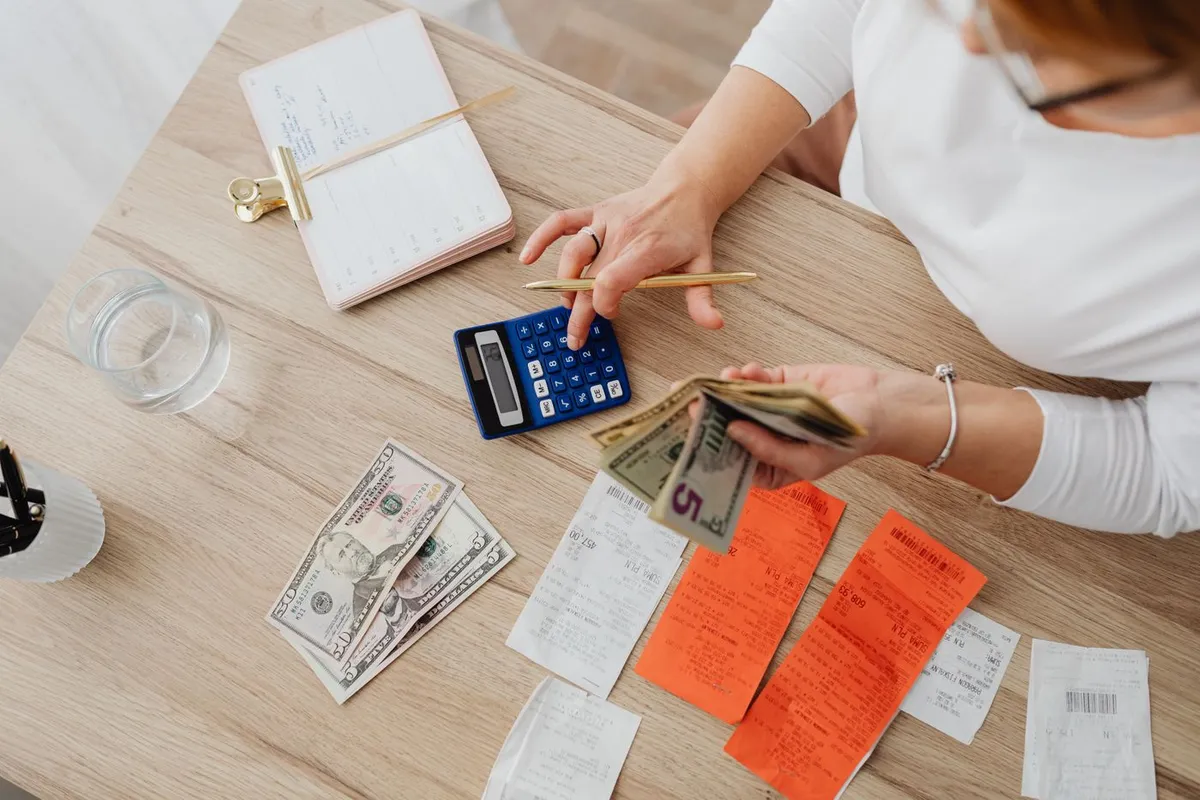 Person using a blue calculator next to dollar bills, receipts and a notebook on a wooden desk