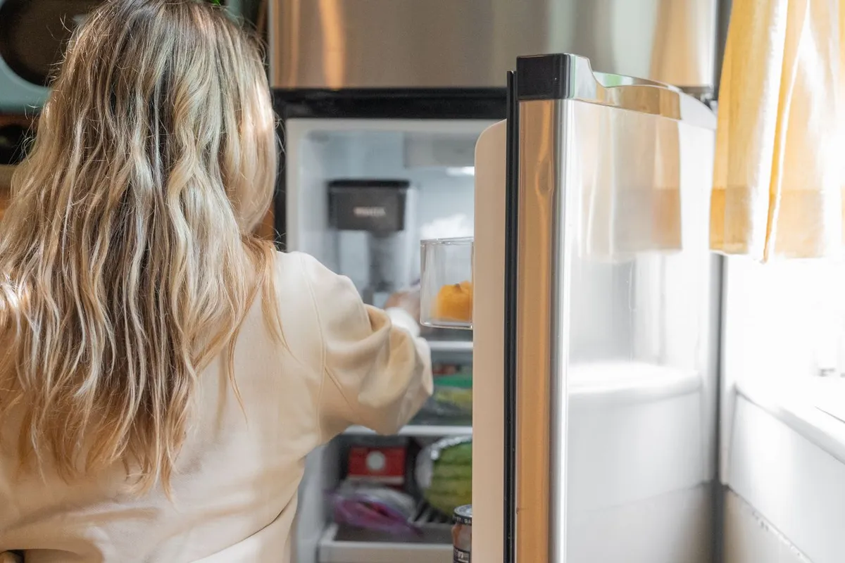 Person with long blonde hair reaching into an open stainless steel refrigerator in a bright kitchen