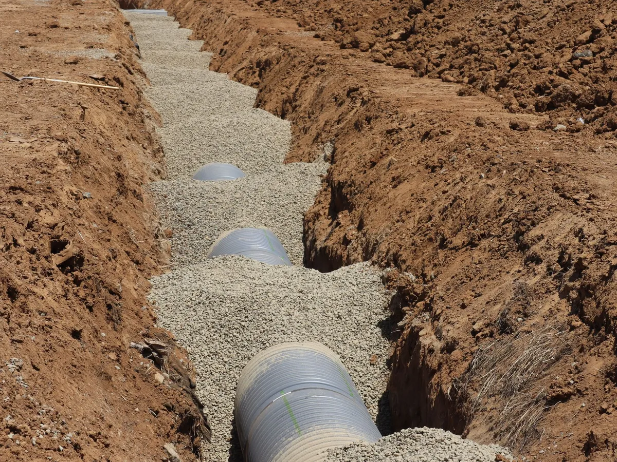 Open trench in red clay soil with ribbed sewer pipe sections laid on a bed of gravel