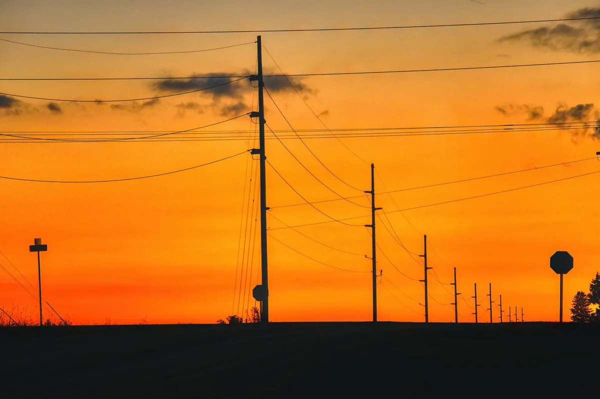 Silhouetted utility poles and power lines against an orange sunset sky in Plainview, Minnesota