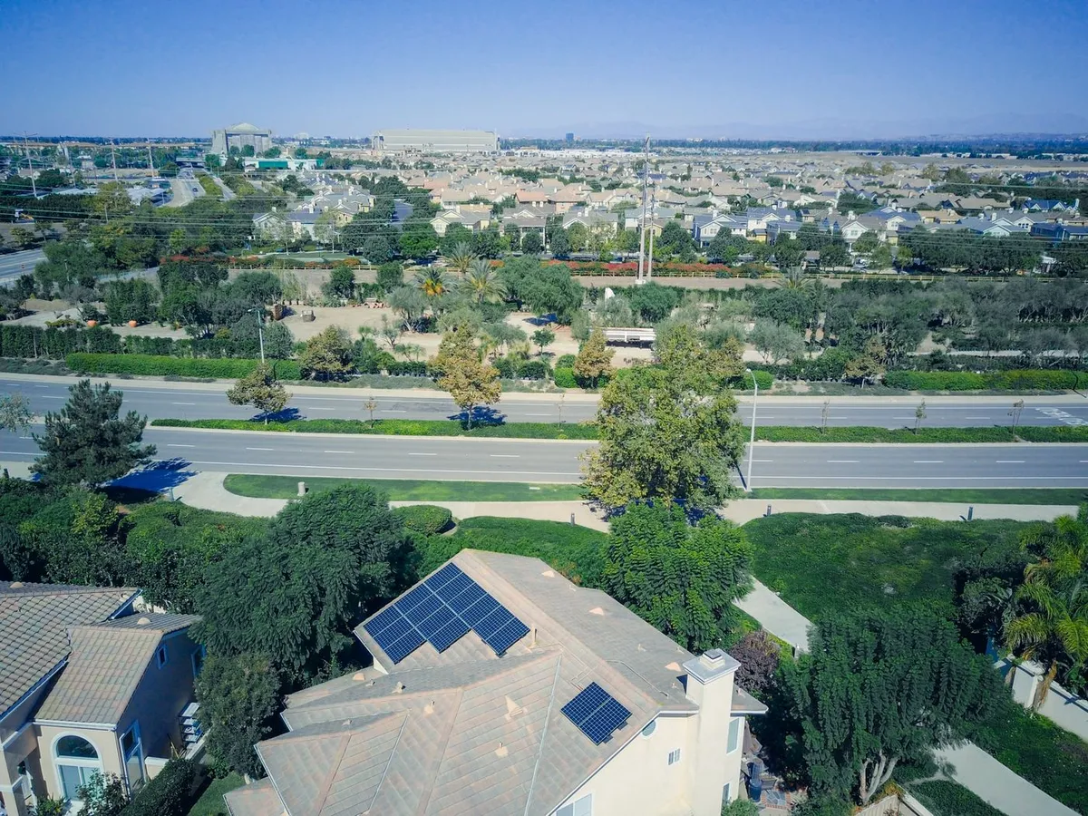 Aerial view of a suburban neighborhood with a house in the foreground that has blue solar panels on its roof, trees and a wide road behind it