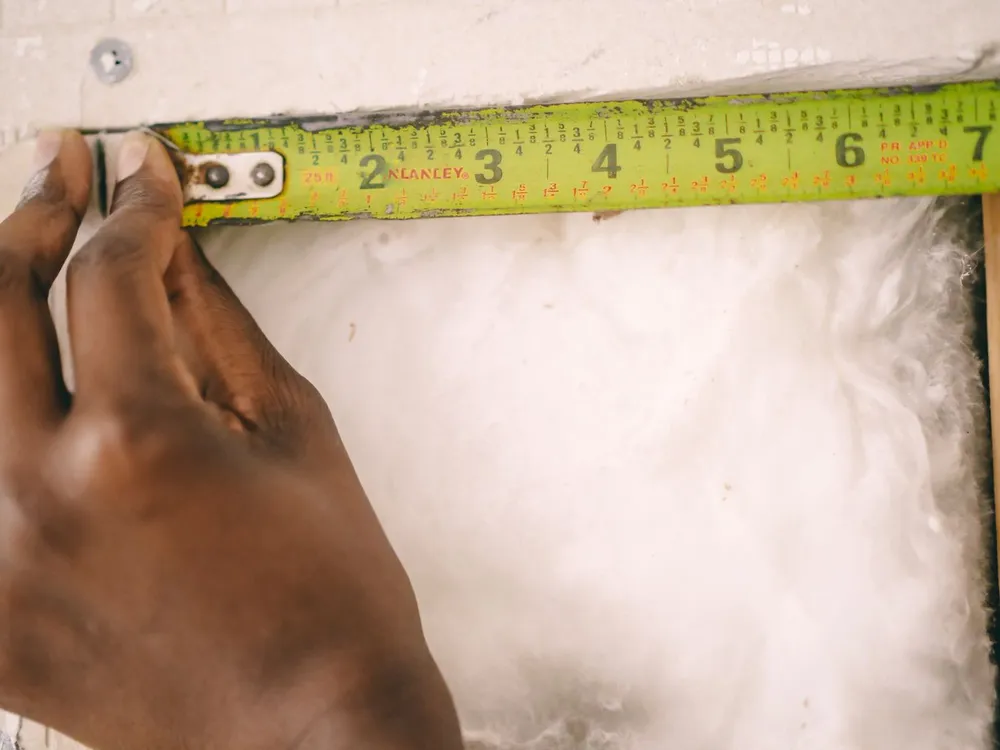 Hand holding a yellow tape measure against fluffy white insulation to read its depth in inches