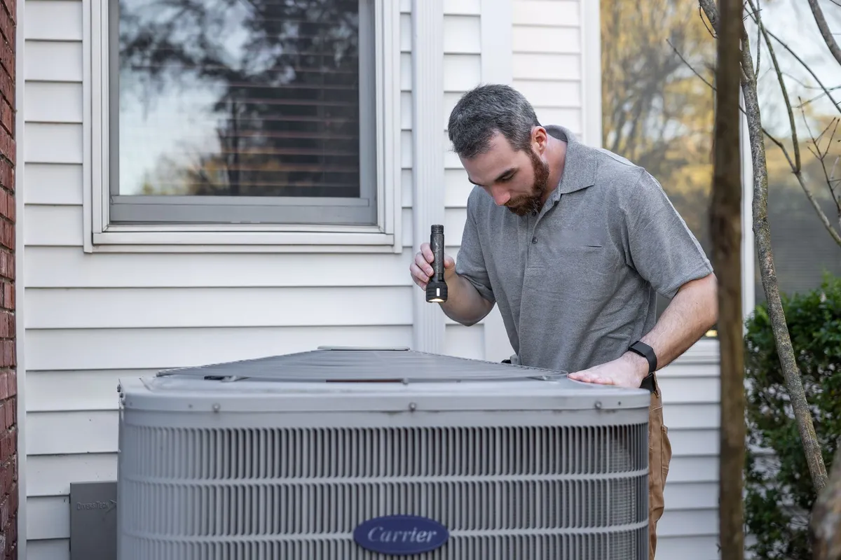 Technician with a flashlight inspecting the top of an outdoor central air conditioner condenser beside a white house