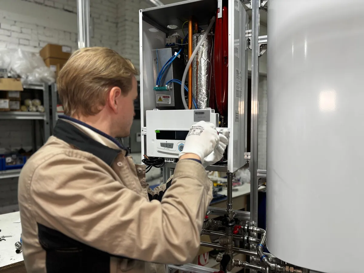 Technician in work gloves adjusting the controls of a wall-mounted heater next to a large white hot water storage tank