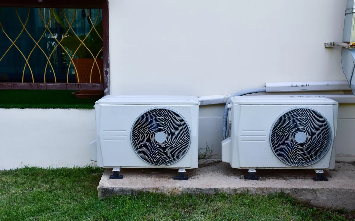 Two white outdoor units standing side by side on a concrete pad against a cream house wall, with grass in front
