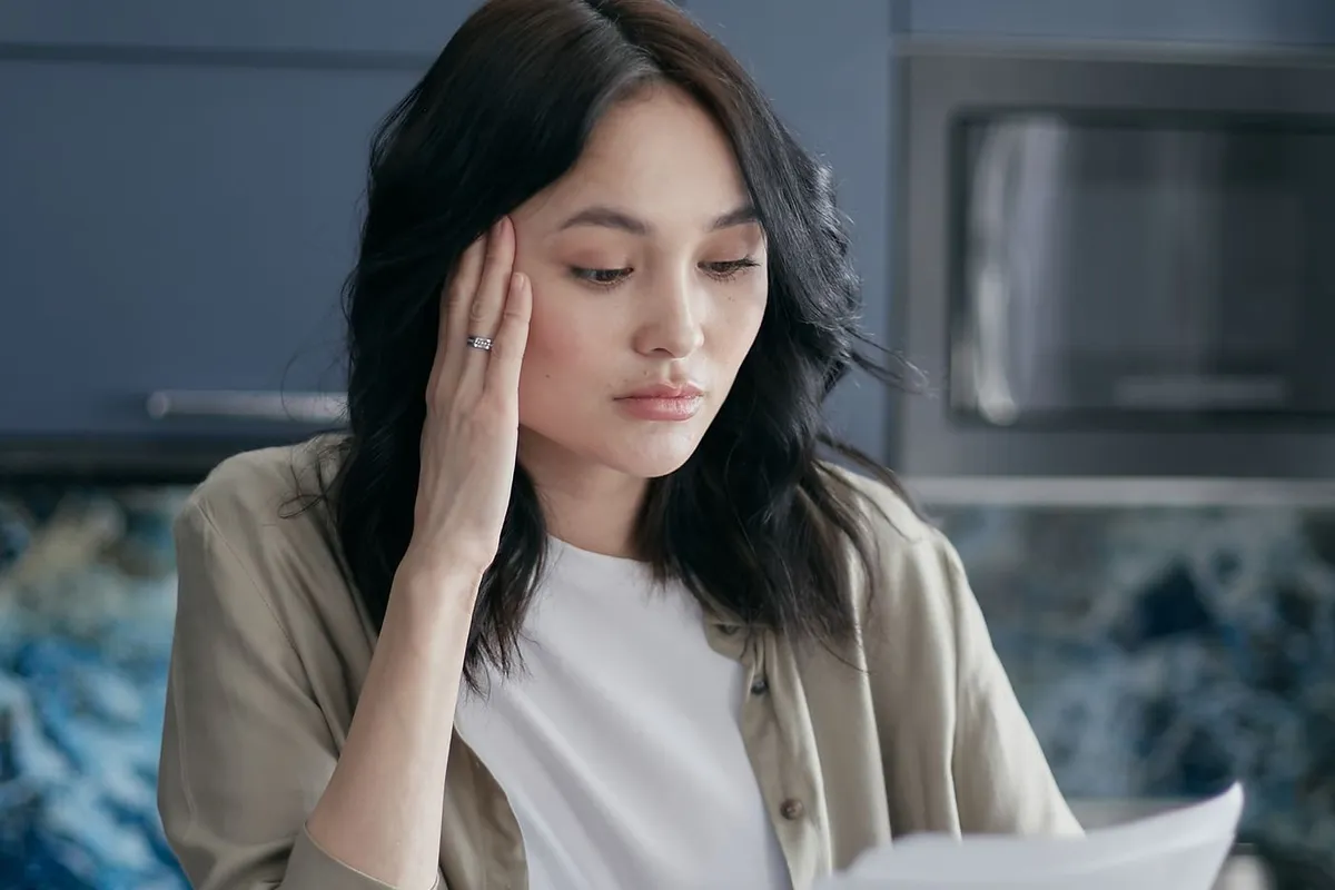 Woman resting her head on one hand while reading a paper bill at a kitchen table, with a calculator and phone in front of her